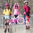 Three children pose for a photo as wear helmets and protective gear for wheeled sports. 